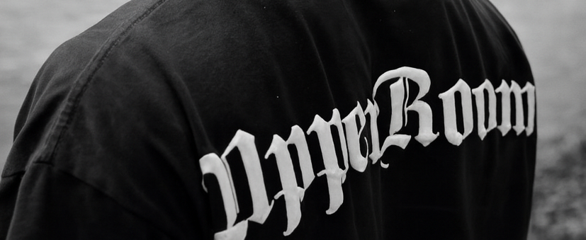 Person wearing a cap and 'Upper Room' t-shirt looking at a lake with mountains in the background